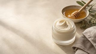 Frosted jar of whipped colostrum tallow balm beside raw honey and linen on a pale stone surface
