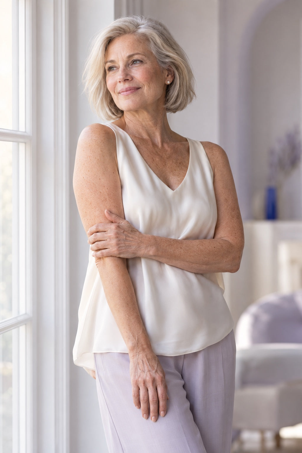 A woman in her sixties in a sleeveless ivory top, standing by a sunlit window